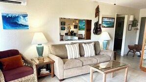 A living room with a beige sofa, a wicker chair, a glass coffee table, and a dining table. There are two lamps on side tables, a guitar on the wall, and a large painting of a wave. - Plantation Hale Suites F7, Ground Floor, AC, Walk to Beach (Kapaa)
