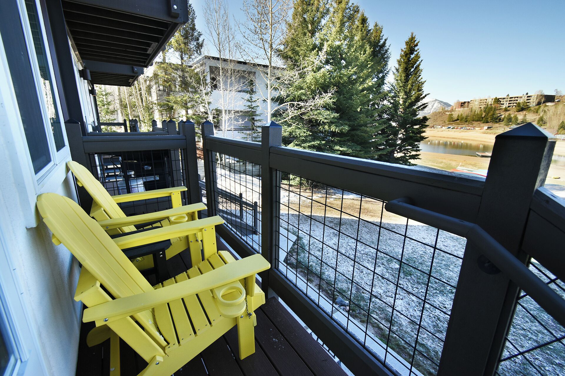 A balcony with yellow Adirondack chairs, a metal railing, and a view of a lake and trees.