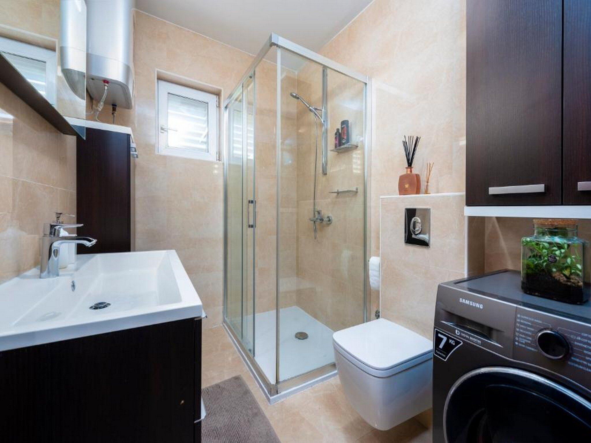 A modern bathroom with a glass shower enclosure, a white sink, a toilet, and a Samsung washing machine.