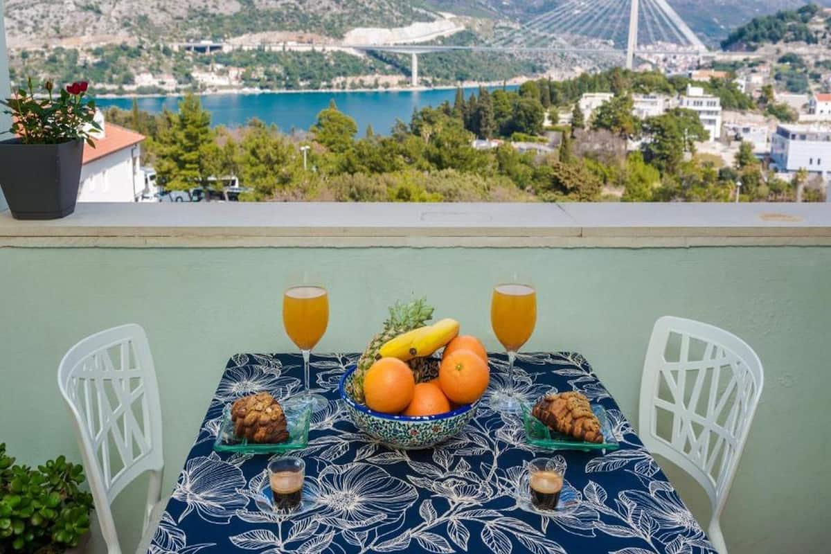 Ein Tisch mit einer blumigen Tischdecke, zwei weiße Stühle, eine Fruchtschale, zwei Gläser Orangensaft und zwei kleine Teller mit Gebäck.
