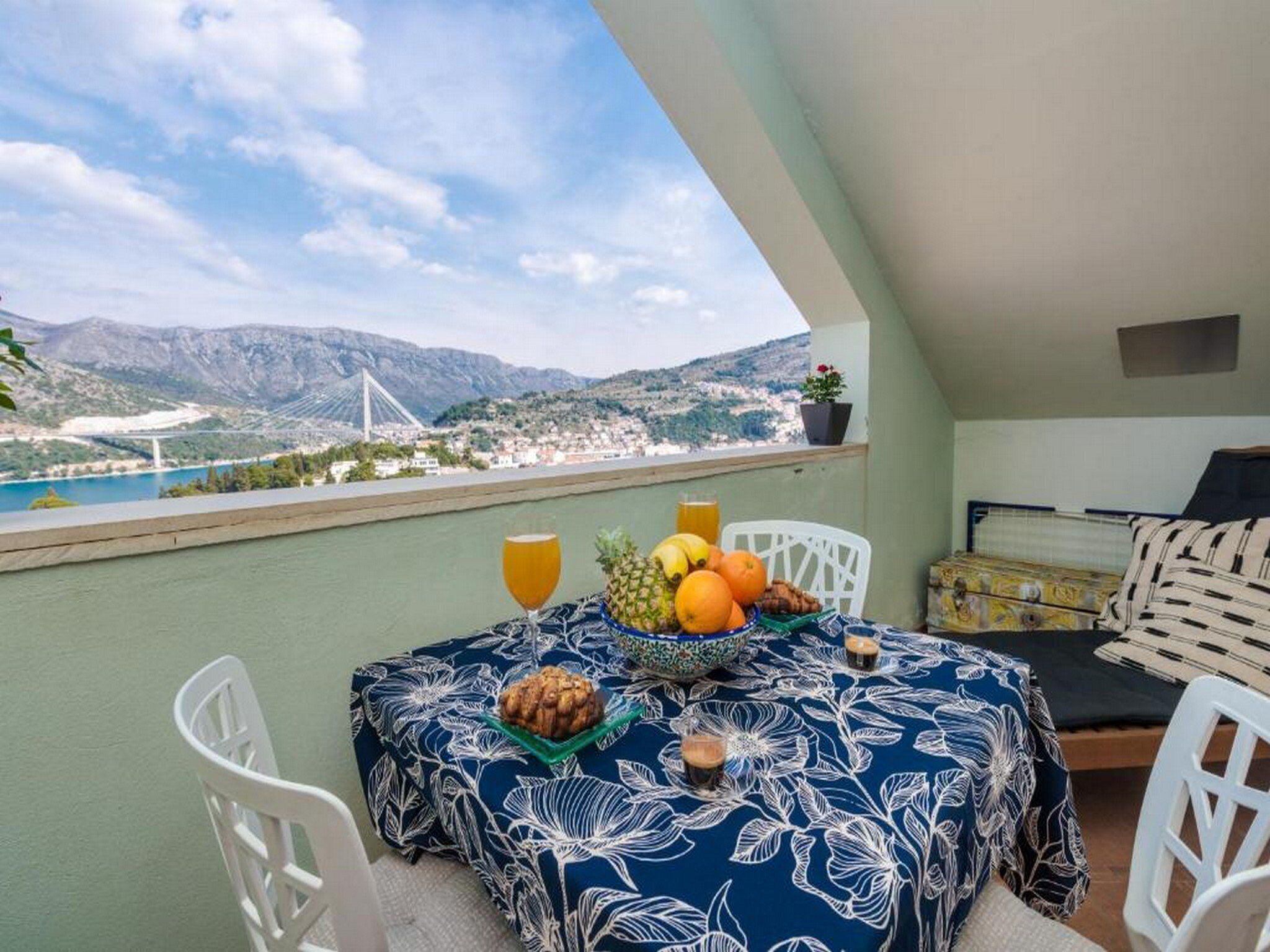 A balcony with a table set for two, featuring a floral tablecloth, a fruit bowl, and two glasses of orange juice.