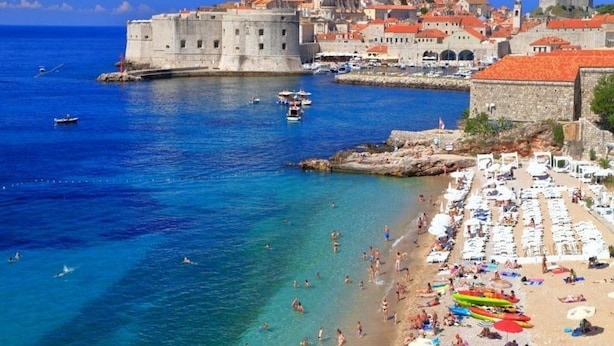 A coastal town with a historic fort, clear blue waters, and a crowded beach with sun loungers. - Apartments Meri & Karla (Dubrovnik)