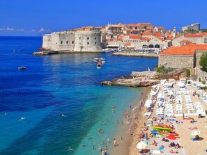 A coastal town with a historic fort, clear blue waters, and a crowded beach with sun loungers. - Apartments Meri & Karla (Dubrovnik)