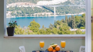 A table with a fruit bowl, two glasses of orange juice, and a pastry, overlooking a lake and a bridge. - Apartments Meri & Karla (Dubrovnik)
