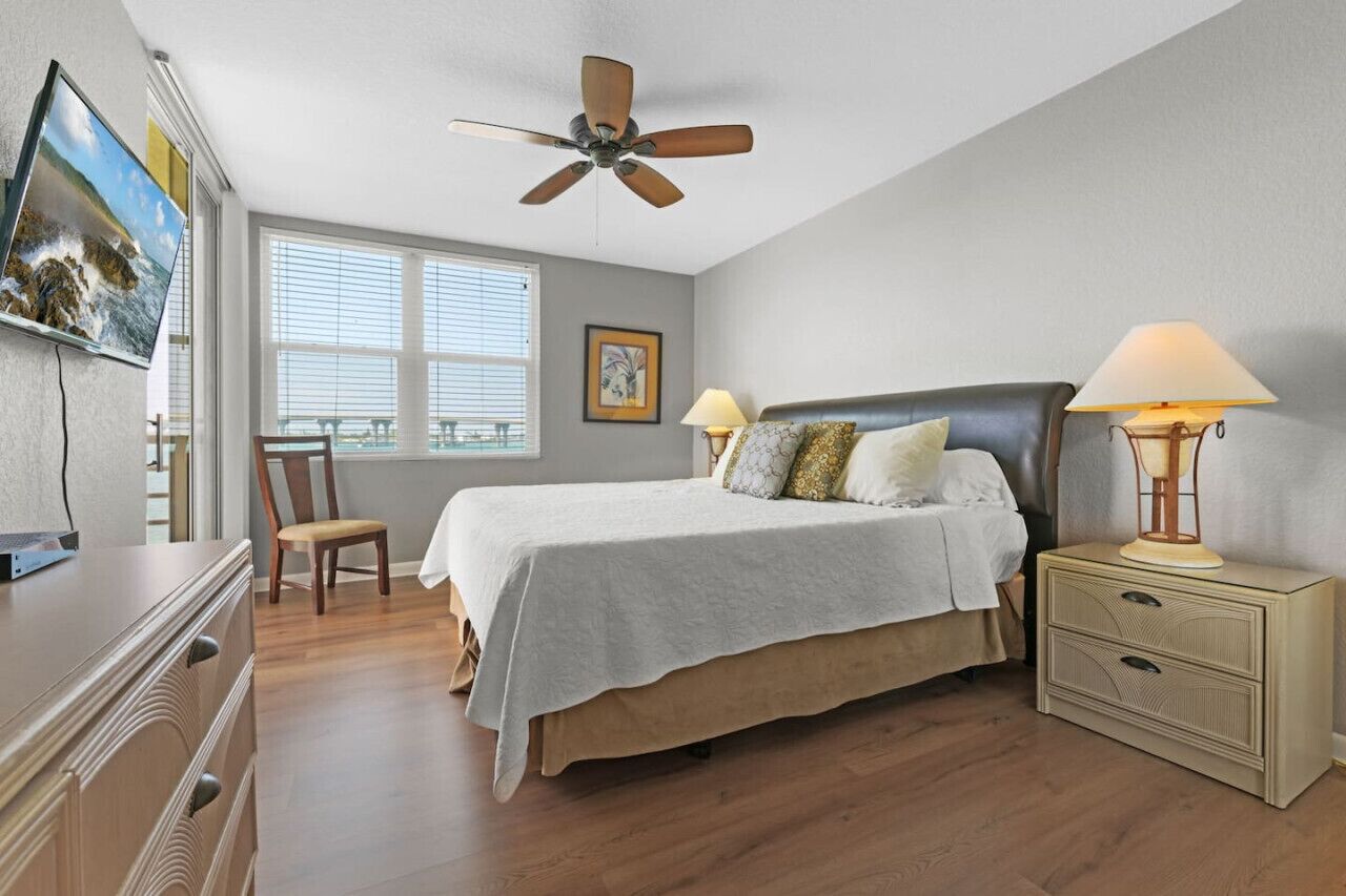 A bedroom with a bed, two bedside lamps, a ceiling fan, a TV mounted on the wall, a chair, and a window with blinds.
