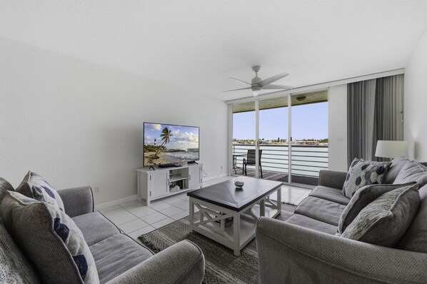 A modern living room with a flat-screen TV, a ceiling fan, and a balcony with a view of the sea.