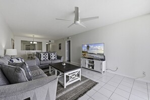 Un salon moderne avec un canapé gris, une table basse, un téléviseur à écran plat posé sur un meuble blanc et un ventilateur de plafond.