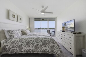 A bedroom with a bed, a ceiling fan, a TV, a dresser, and a balcony with a view of the sea.