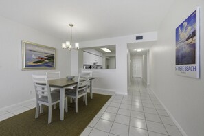 A dining area with a table and chairs, a kitchen in the background, and a hallway leading to a door.