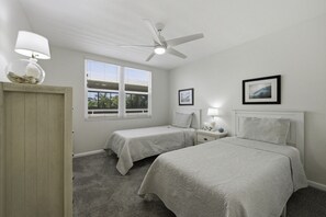 A bedroom with two beds, a ceiling fan, a window with blinds, and a nightstand with a lamp.
