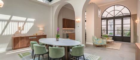 A spacious dining area with a round table and green chairs, a kitchen in the background, and a large arched window with a view.