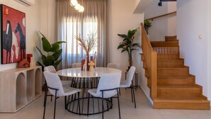 A dining area with a round table, six chairs, and a chandelier. There is a staircase with wooden steps and a potted plant on the landing.