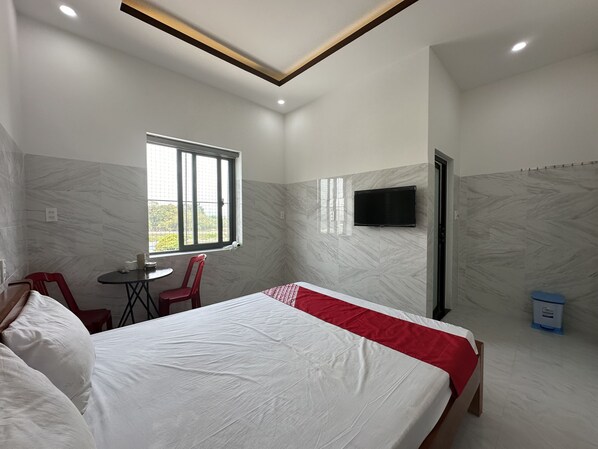 Quarto de hotel com cama, televisão na parede, janela e uma pequena mesa com cadeiras.
