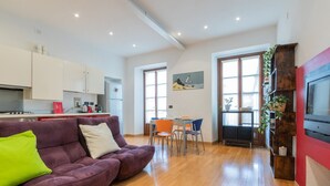 A modern apartment with a living room featuring a purple sofa, a dining area with a table and chairs, a kitchen with white cabinets, and a TV mounted on a red wall. - La Corte by Wonderful Italy (Torino)