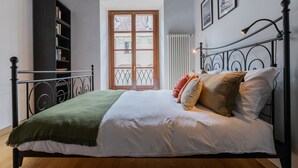 A neatly made bed with a green throw blanket, a white comforter, and several pillows. There is a bookshelf to the left and a window with a view of buildings outside. - La Corte by Wonderful Italy (Torino)