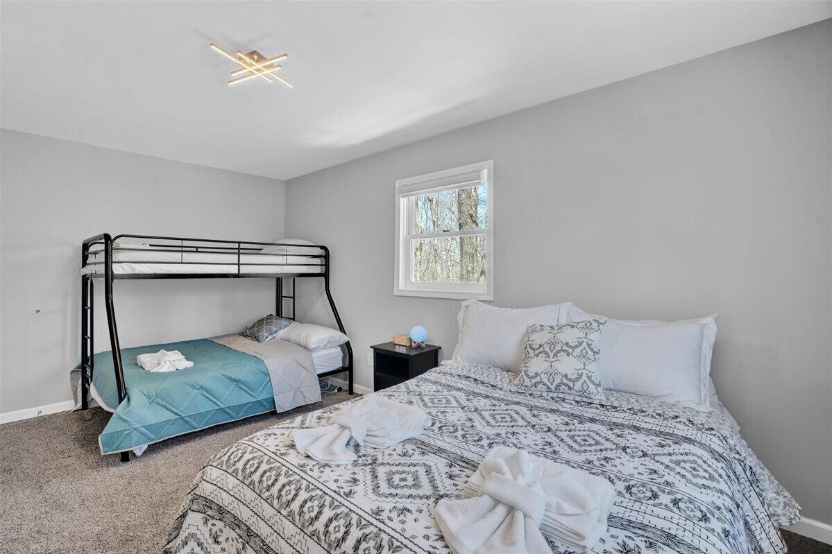 A bedroom with a bunk bed, a single bed, a nightstand, a lamp, and a window with a view of trees.