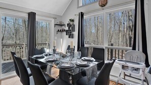 A dining area with a glass table set for six, black chairs, and a high chair. Large windows offer a view of trees. - Spacious and Remodeled Cabin in Poconos Mountain Resort. Hot Tub and Game Room! (Clifton Township)