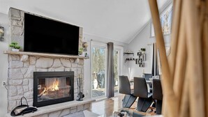 A modern living room with a stone fireplace, a flat-screen TV, and a dining area with chairs and a table. - Spacious and Remodeled Cabin in Poconos Mountain Resort. Hot Tub and Game Room! (Clifton Township)
