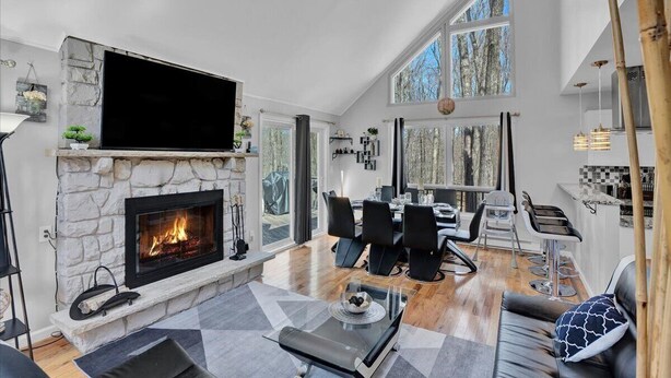 A modern living room with a stone fireplace, a flat-screen TV, and a dining area with black chairs and a glass table. - Spacious and Remodeled Cabin in Poconos Mountain Resort. Hot Tub and Game Room! (Clifton Township)
