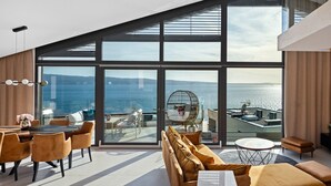 Een modern interieur met grote ramen met uitzicht op zee. Er is een eethoek met een tafel en stoelen, een zithoek met banken en een ronde tafel.