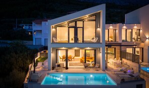 Une maison moderne à deux étages, dotée d’une piscine, d’un espace salon extérieur et de grandes fenêtres.