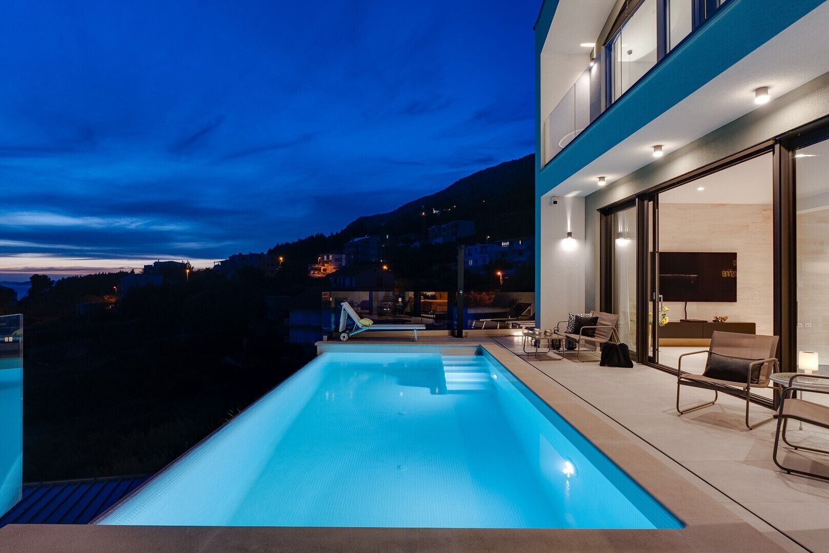 Un área de piscina moderna al aire libre con cerramiento de cristal, sillas de descanso y vistas del paisaje circundante.