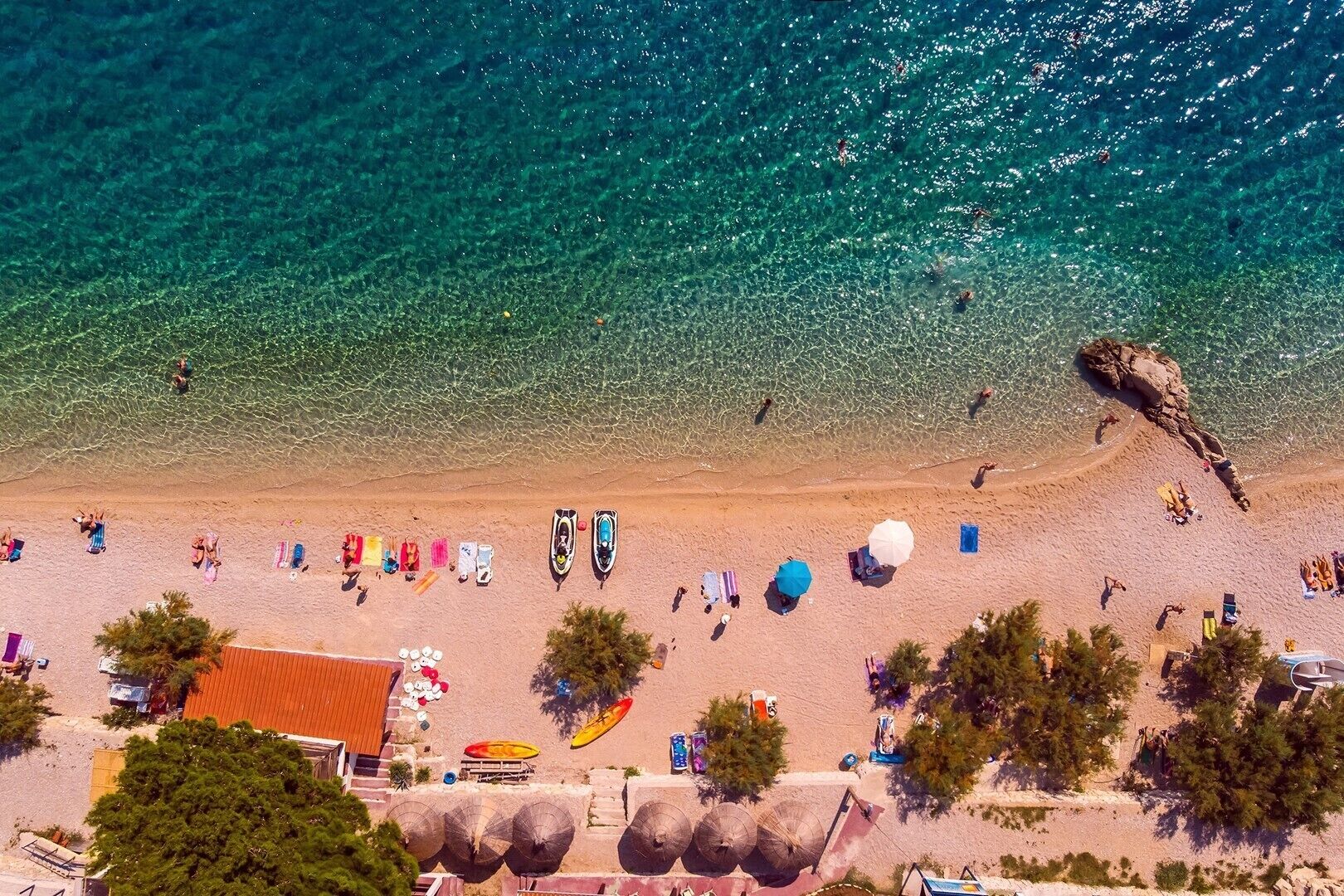 An aerial view of a beach with clear turquoise waters, a sandy shore, and various beach activities.