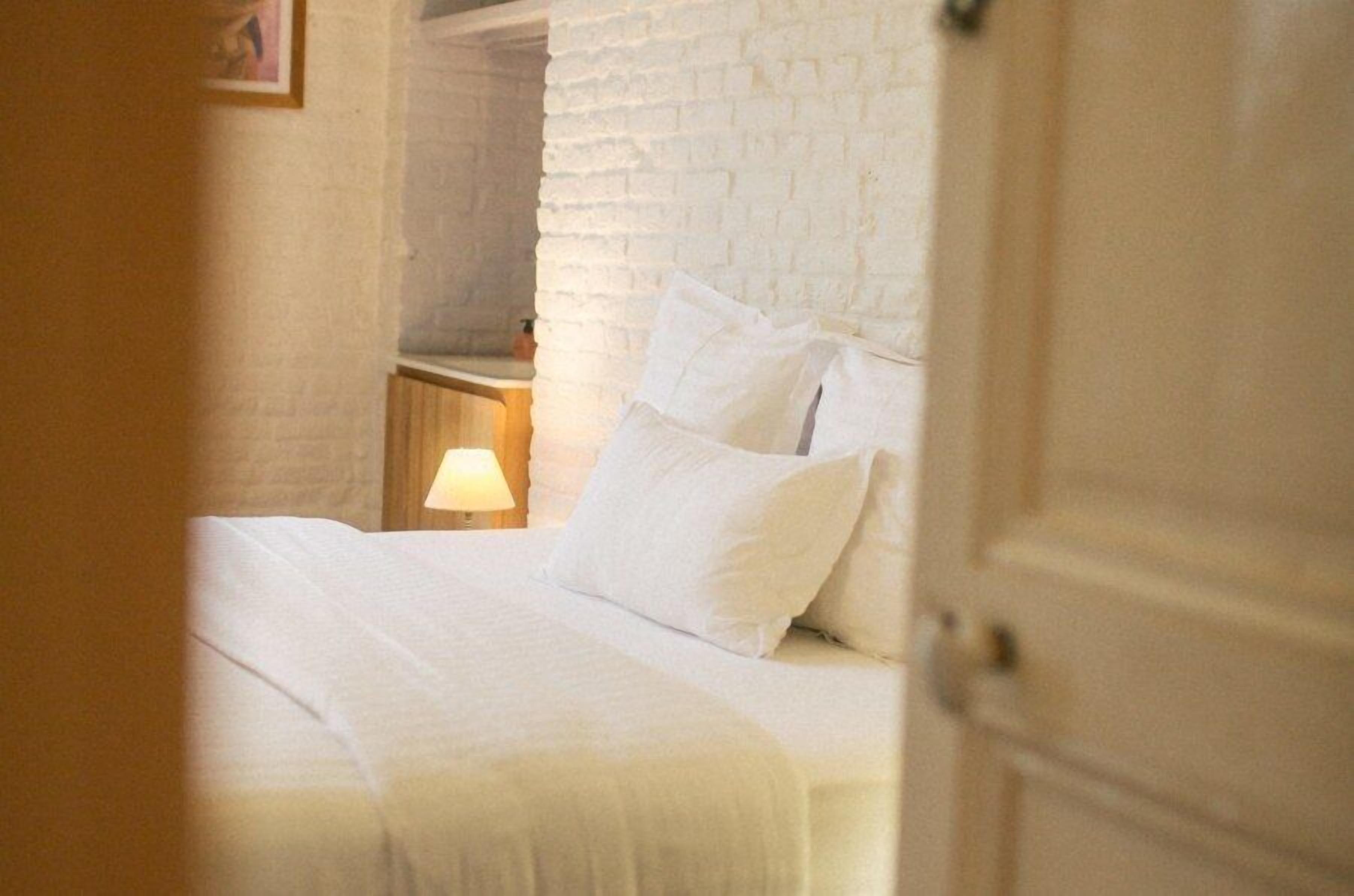 Un dormitorio con pared de ladrillo blanco, una cama con ropa de cama blanca y una mesita de noche con lámpara.