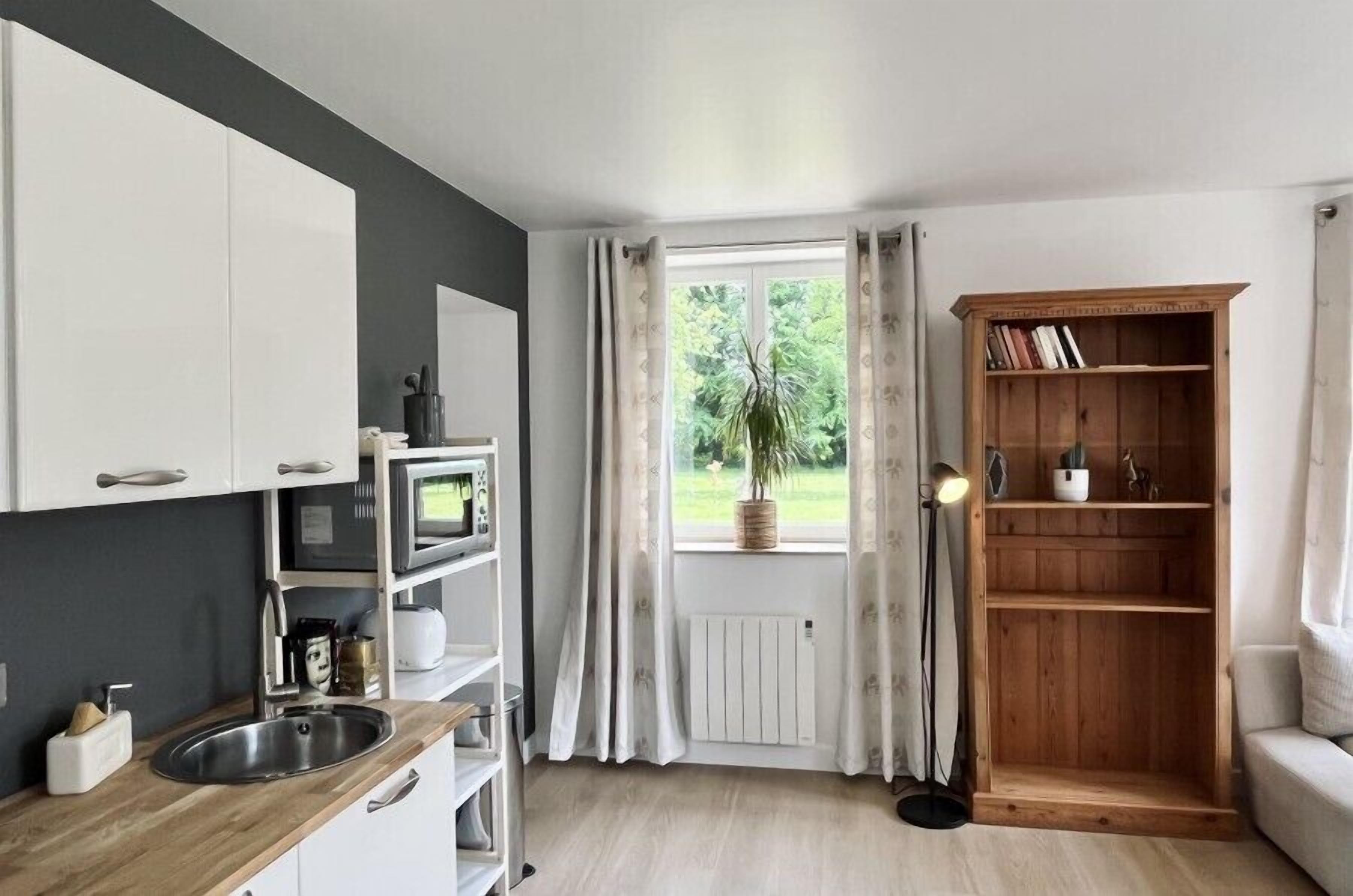 Une cuisine de taille réduite avec des meubles blancs, un four à micro-ondes et un évier. On y trouve une fenêtre avec des rideaux, une bibliothèque et un lampadaire.