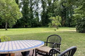 Een ronde tafel met een tafelkleed met patroon, twee zwarte rieten stoelen en een gazon met bomen op de achtergrond.