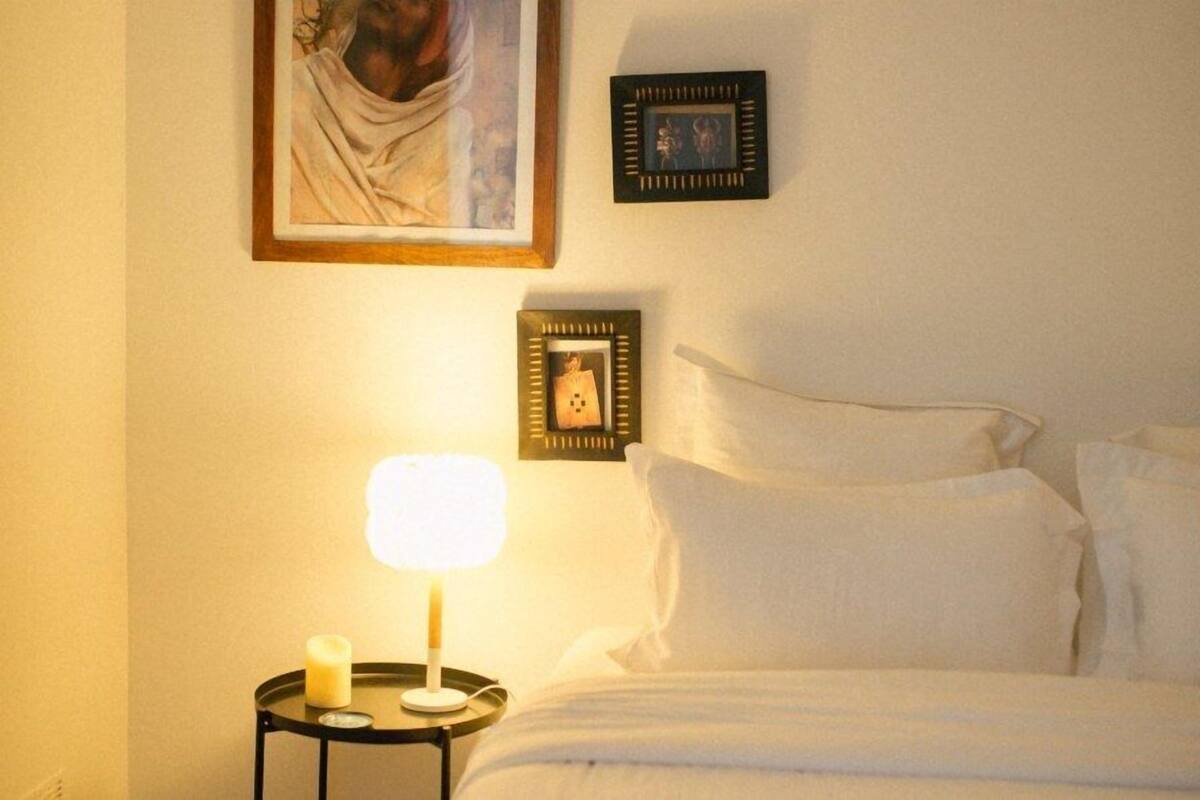 Ein ordentlich gemachtes Bett mit weißen Leinen, ein Nachttisch mit Lampe und Kerze sowie gerahmte Bilder an der Wand.