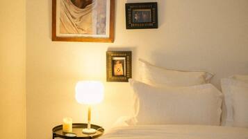 A neatly made bed with white linens, a bedside table with a lamp and a candle, and framed pictures on the wall.