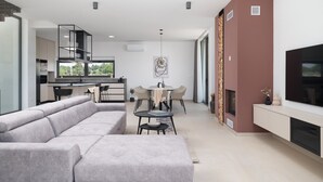 A modern living room with a grey sectional sofa, a dining area with a round table and chairs, a kitchen with a built-in stove, and a TV mounted on the wall. - New Villa 55-Vrsi heated pool jacuzzi sauna bbq sea view (Vrsi)