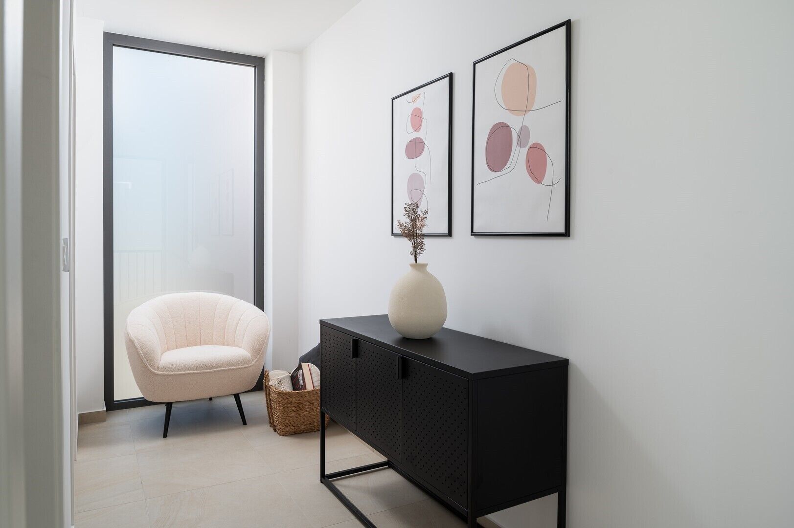 Una stanza moderna con una porta scorrevole in vetro che dà su un balcone, una poltrona color crema, un tavolino consolle nero con un vaso e opere d'arte astratte alle pareti.