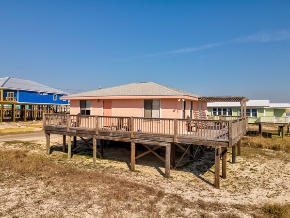 A beachfront house with a wooden deck and a small porch, surrounded by sand and sparse vegetation.