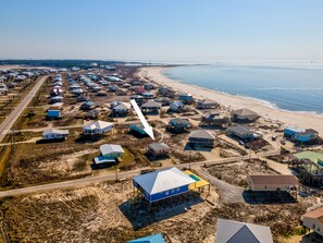 Vue aérienne d’un quartier résidentiel situé sur la côte, avec des maisons, dont certaines sont surélevées sur des piliers, et une plage en arrière-plan.