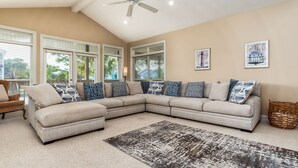 A spacious living room with a large L-shaped sectional sofa, a ceiling fan, and a large window with blinds. - Another Happy Clam, Beachside, 4/2, community POOL, dog friendly, sleeps 10 (Dauphin Island)