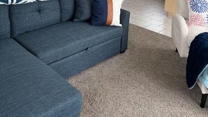 Een moderne woonkamer met een hoekbank, een eethoek en een keuken met houten kastjes en een zwarte koelkast.