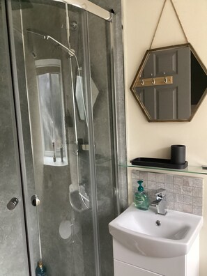 Een badkamer met een glazen douchecabine, een wit wastafeltje, een spiegel en een zeepdispenser.