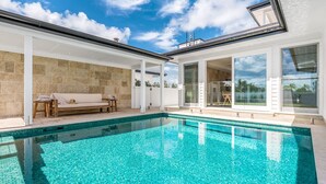 Un área de piscina exterior moderna con un muro de piedra, un banco de madera y una puerta de vidrio que da a un porche cubierto.
