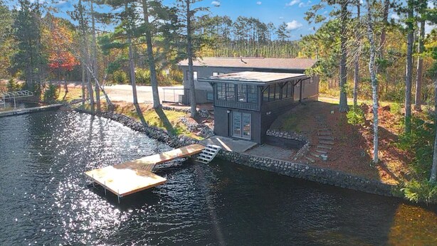 Ein modernes Haus mit Steg, der sich in ruhiges Wasser erstreckt, umgeben von Bäumen und blauem Himmel.