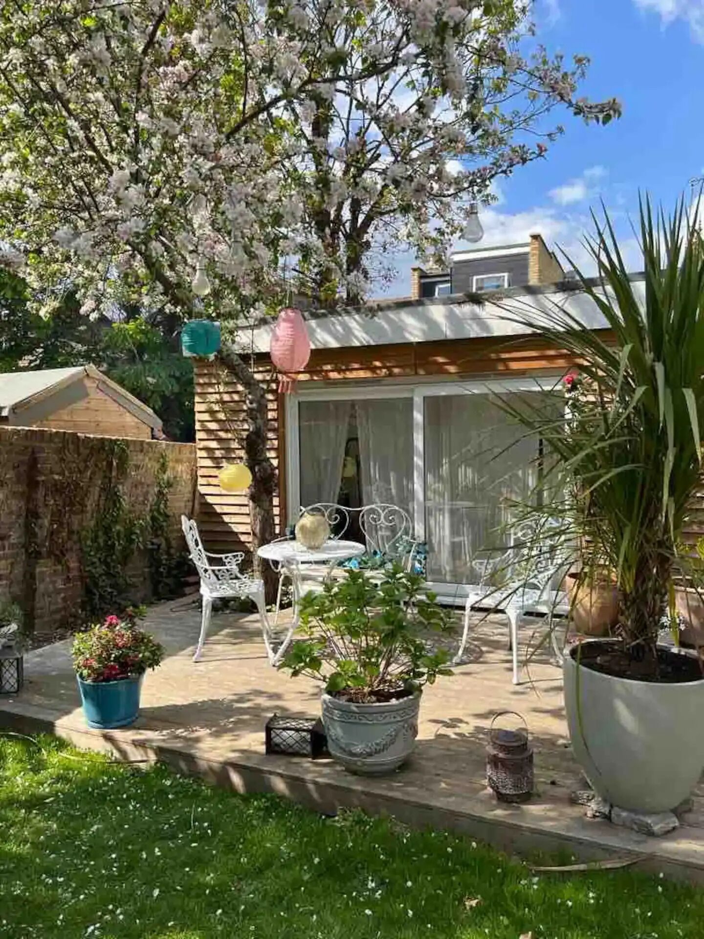 Un patio con mesa y sillas, rodeado de plantas en macetas y un gran árbol con flores.
