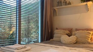 Ein ordentlich bezogenes Bett mit gefalteten Handtüchern, ein großes Fenster mit Jalousien und ein Regal mit Dekoration.