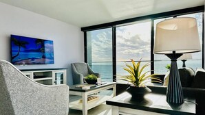 Una sala moderna con un gran ventanal con vista al océano, un televisor de pantalla plana, un sillón gris, una mesita con una lámpara y una planta en maceta.