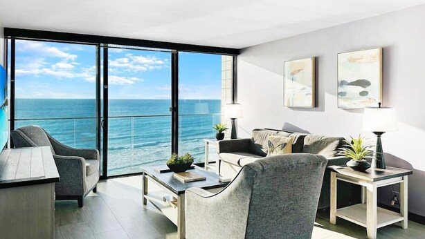 Una sala de estar moderna con un amplio ventanal corredera que da a un balcón con vistas al océano. Hay dos sofás, una mesa de centro y una mesita auxiliar con una lámpara.
