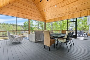 A covered outdoor deck with a table and chairs, large windows, and a view of a forest.