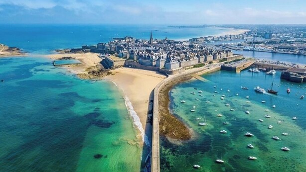 Vista aérea de una ciudad costera con un centro histórico fortificado, una playa de arena y un puerto deportivo con numerosos barcos.