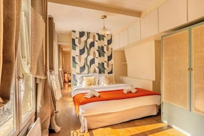 A hotel room with a bed, a large window with curtains, a patterned wall, and a rattan wardrobe.