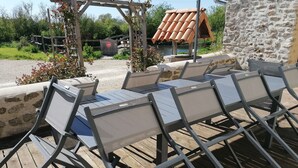 A wooden deck with outdoor seating, including tables and chairs, and a stone wall. - Gîte à la Campagne, 3km de Parthenay. Idéal Pour Séjours en Famille Entre Amis (PARTHENAY)
