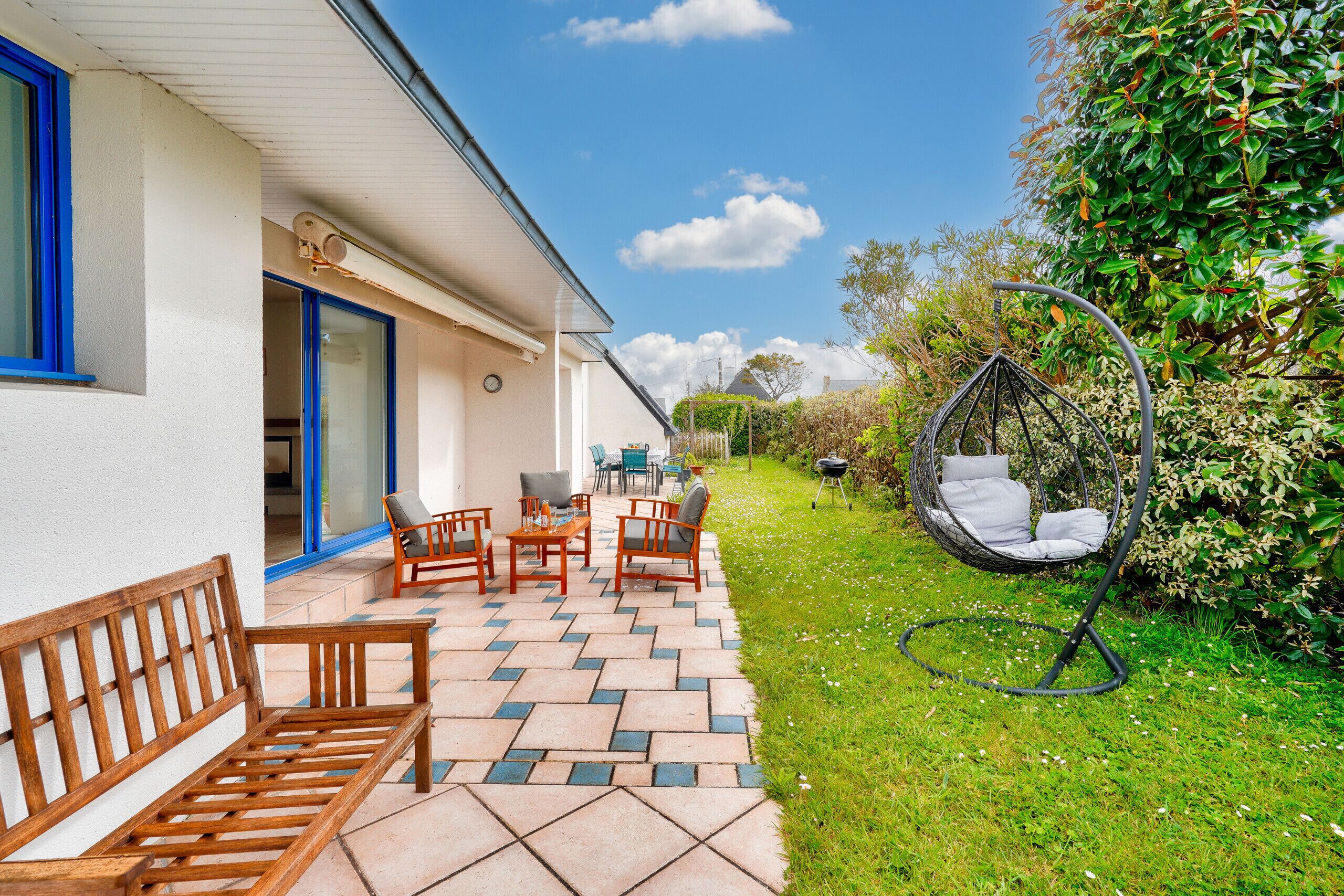 Un patio con un banco de madera, una mesa y sillas, una silla colgante y un jardín con seto y columpio.